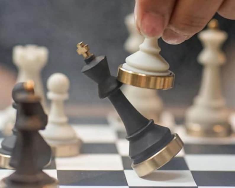 Op de afbeelding: Een zwarte schaakkoning met gouden accenten wordt omvergeworpen door een wit schaakstuk. De schaakstukken staan op een zwart-wit geblokt bord. Andere schaakstukken zijn op de achtergrond te zien.