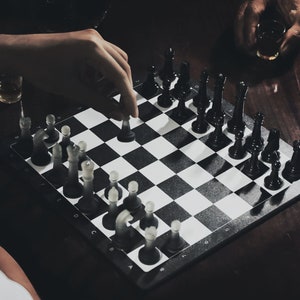 May include: A black and white chessboard with black chess pieces on one side and white chess pieces on the other. A hand is moving a white chess piece.