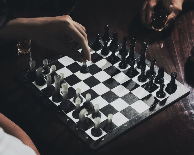 May include: A black and white chessboard with black chess pieces on one side and white chess pieces on the other. A hand is moving a white chess piece.