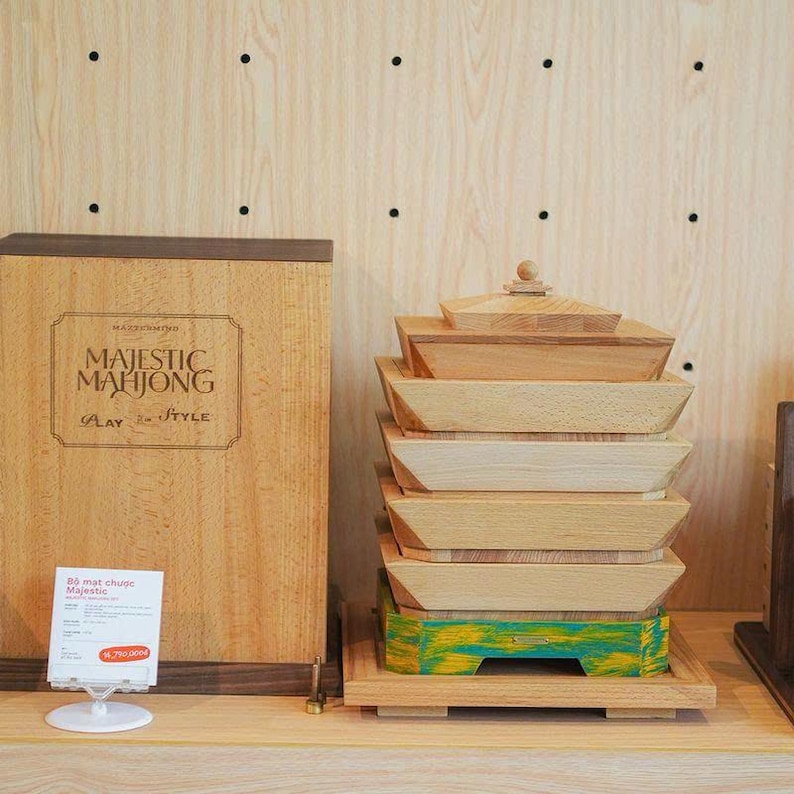 May include: A wooden Majestic Mahjong set with a stack of wooden trays and a box. The box has the words "Majestic Mahjong" printed on it. The trays are stacked on a wooden base with a green and yellow painted bottom.