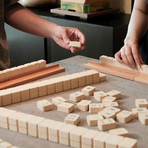 May include: A close-up shot of a Mahjong game in progress. The game features beige tiles with green symbols and numbers, arranged on a gray surface. Wooden racks and a wooden tray are also visible.