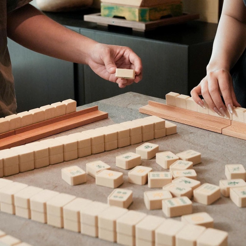 May include: A close-up shot of a Mahjong game in progress. The game features beige tiles with green symbols and numbers, arranged on a gray surface. Wooden racks and a wooden tray are also visible.