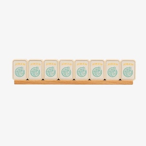 May include: A wooden Mahjong rack holding eight tiles. Each tile is cream-colored with the word "JOKER" in gold and a teal design. The rack is made of light-colored wood.
