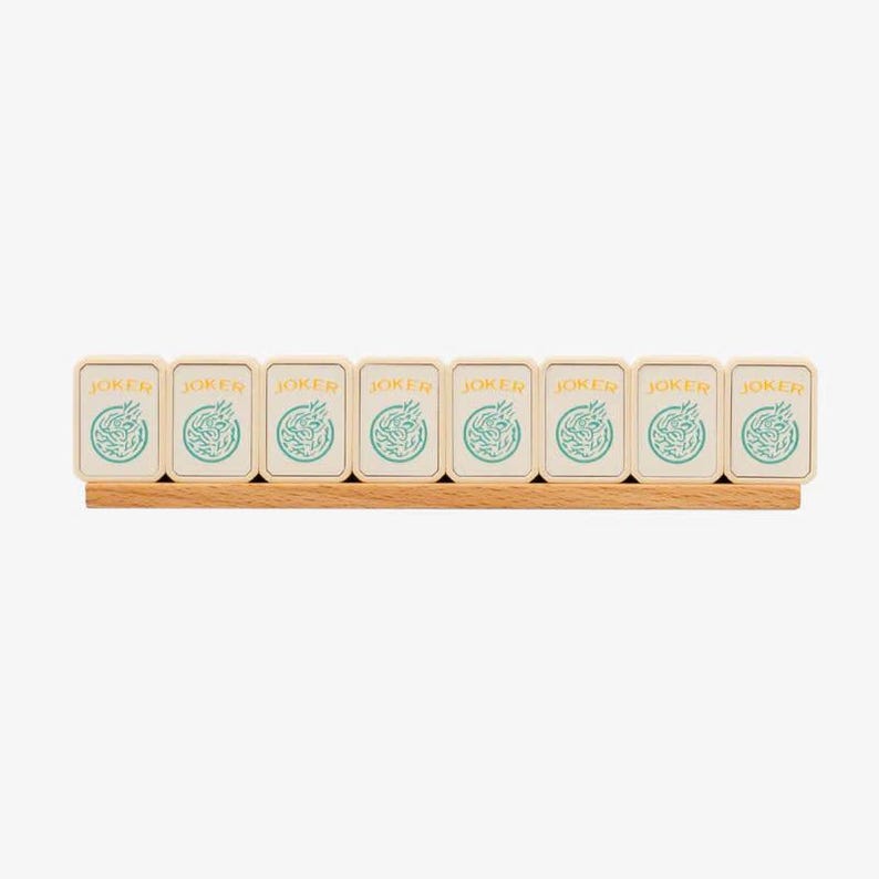 May include: A wooden Mahjong rack holding eight tiles. Each tile is cream-colored with the word "JOKER" in gold and a teal design. The rack is made of light-colored wood.
