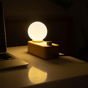 May include: A modern wooden desk lamp with a spherical, glowing light. The lamp sits on a light-colored surface next to a laptop. The lamp has a rectangular wooden base with a small knob and a black power cord.