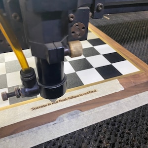 Op de afbeelding: Een lasergraveermachine in werking, gericht op een houten schaakbord. Het bord heeft een zwart-wit geblokt patroon. De tekst "Success is not final, failure is not fatal" is in het hout gegraveerd.