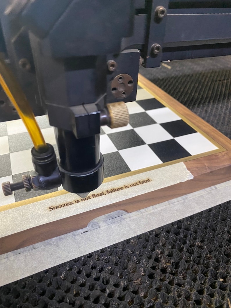 Op de afbeelding: Een lasergraveermachine in werking, gericht op een houten schaakbord. Het bord heeft een zwart-wit geblokt patroon. De tekst "Success is not final, failure is not fatal" is in het hout gegraveerd.