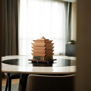 May include: A wooden pagoda-style decorative object with multiple stacked tiers, in shades of brown and tan, sits on a round table. The table has a black and white marble top. A modern chandelier hangs above the table, and a chair is in the foreground.