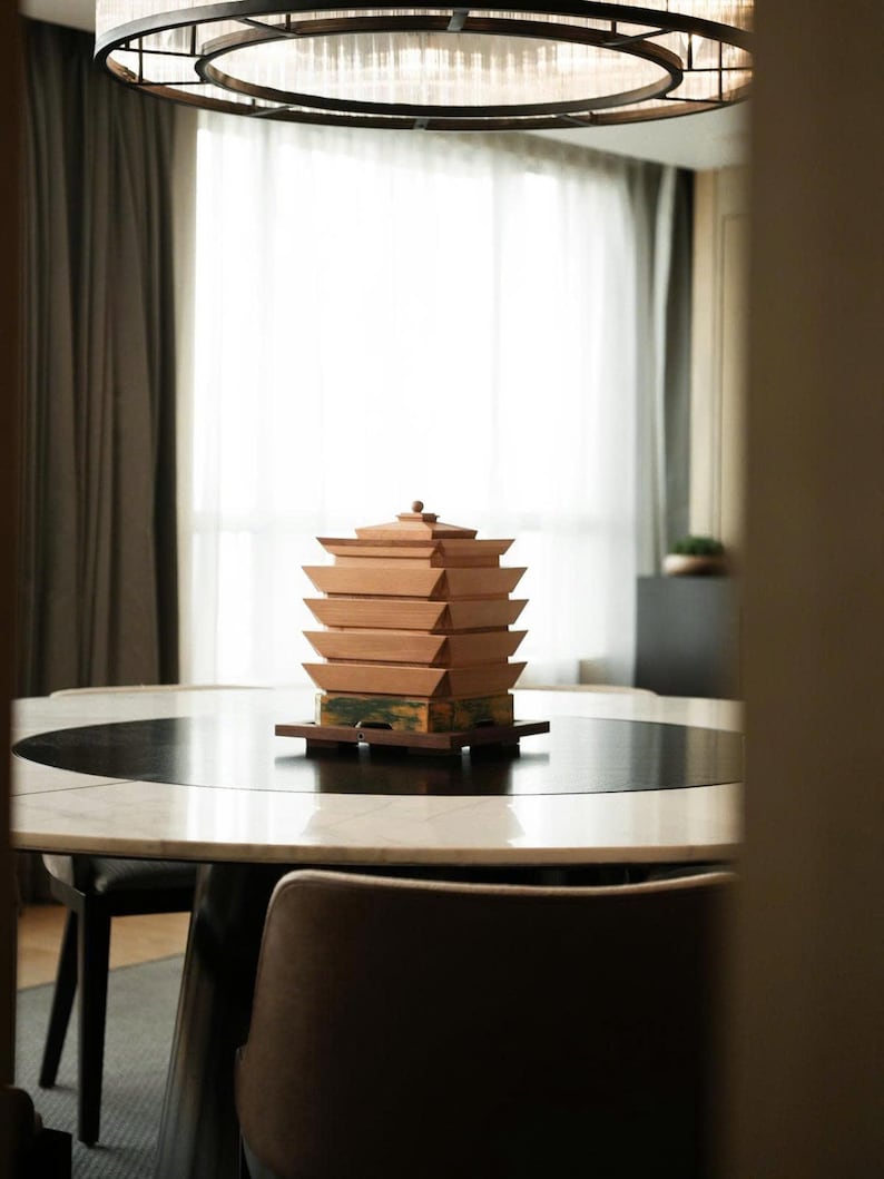 May include: A wooden pagoda-style decorative object with multiple stacked tiers, in shades of brown and tan, sits on a round table. The table has a black and white marble top. A modern chandelier hangs above the table, and a chair is in the foreground.
