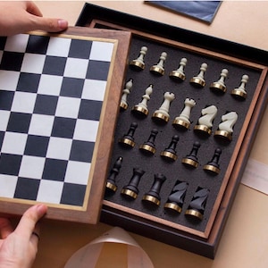 Op de afbeelding: Een houten schaakspel met een zwart-wit geblokt bord en een compartiment met de schaakstukken. De stukken zijn zwart en wit met goudkleurige accenten. De set wordt open gepresenteerd.