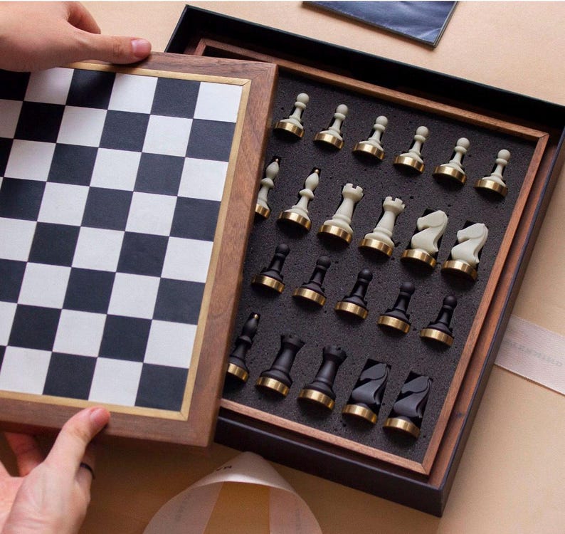 Op de afbeelding: Een houten schaakspel met een zwart-wit geblokt bord en een compartiment met de schaakstukken. De stukken zijn zwart en wit met goudkleurige accenten. De set wordt open gepresenteerd.