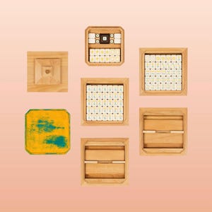 May include: An overhead shot of a collection of wooden game pieces and boxes, likely for a board game. The pieces are arranged in a circular pattern on a light pink background. The boxes are square and rectangular, some containing tiles with symbols.