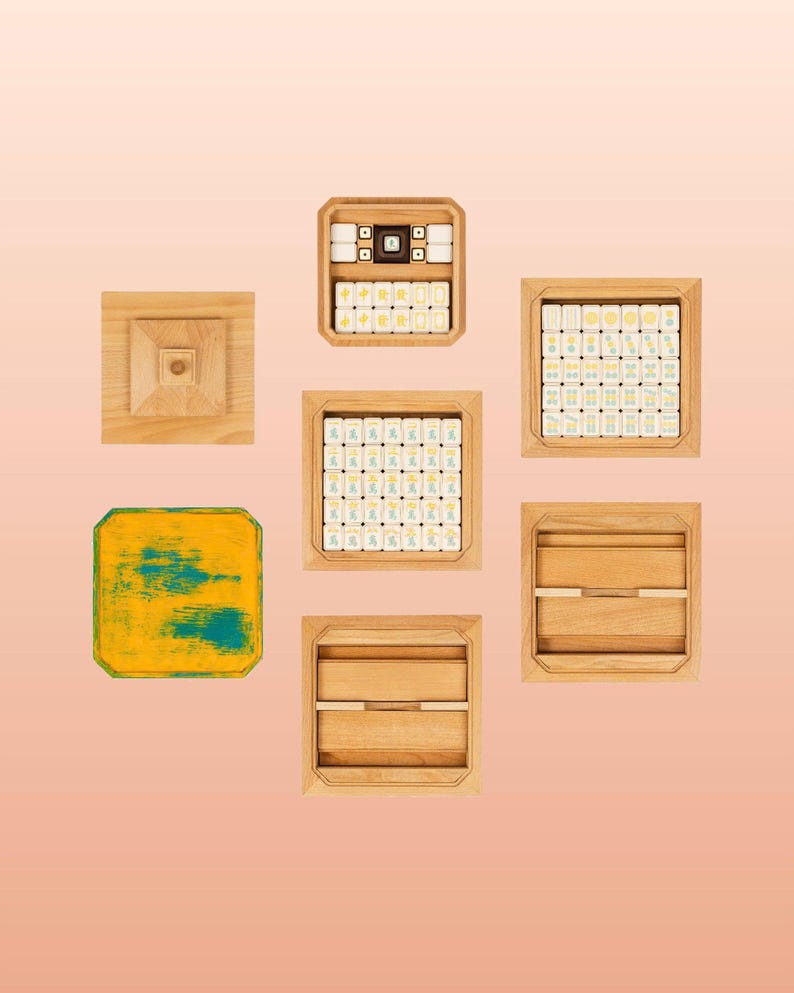 May include: An overhead shot of a collection of wooden game pieces and boxes, likely for a board game. The pieces are arranged in a circular pattern on a light pink background. The boxes are square and rectangular, some containing tiles with symbols.