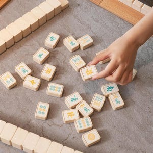 May include: A close-up shot of a Mahjong set with beige tiles and wooden racks. The tiles feature Chinese characters and symbols in green and yellow. A hand is seen selecting a tile from the scattered pieces.