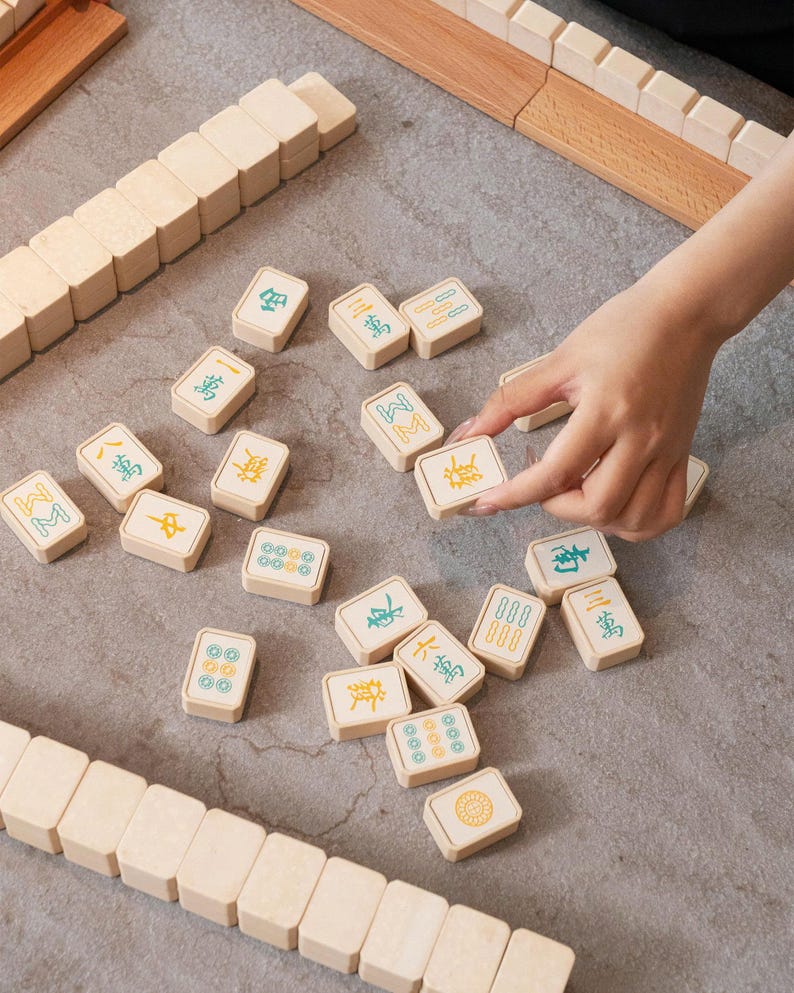May include: A close-up shot of a Mahjong set with beige tiles and wooden racks. The tiles feature Chinese characters and symbols in green and yellow. A hand is seen selecting a tile from the scattered pieces.