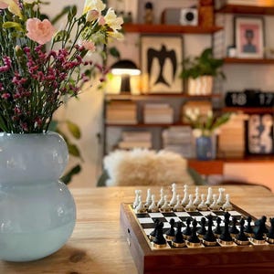 Op de afbeelding: Een houten schaakspel met zwarte en witte stukken staat op een houten tafel. Het schaakbord is opgesteld met alle stukken in hun startposities. Een vaas met bloemen staat op de voorgrond.