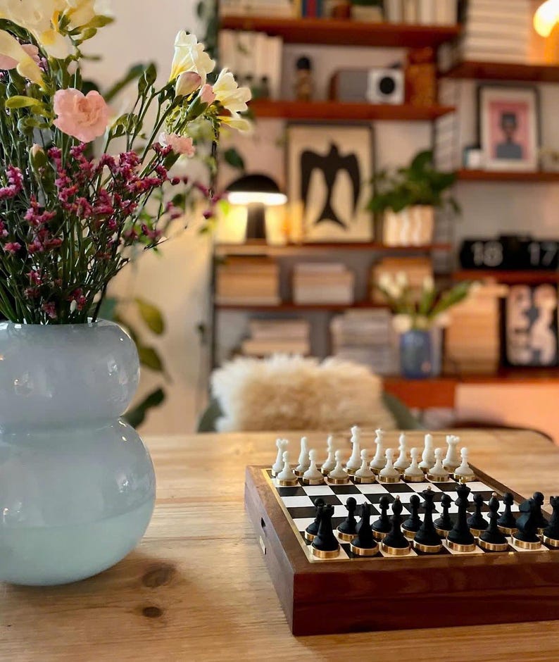 Op de afbeelding: Een houten schaakspel met zwarte en witte stukken staat op een houten tafel. Het schaakbord is opgesteld met alle stukken in hun startposities. Een vaas met bloemen staat op de voorgrond.