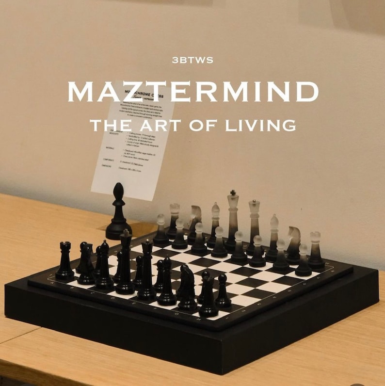 May include: A black and white chess set on a black chessboard with a black wooden frame. The chessboard is on a wooden table. The text "MAZTTERMIND THE ART OF LIVING" is visible above the chessboard.