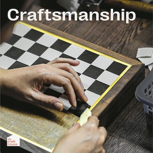 Op de afbeelding: Een close-up van een schaakbord met zwart-witte vierkanten, omlijst door donker hout met een gouden rand. Het woord "Craftsmanship" staat bovenaan. Handen werken aan het bord.