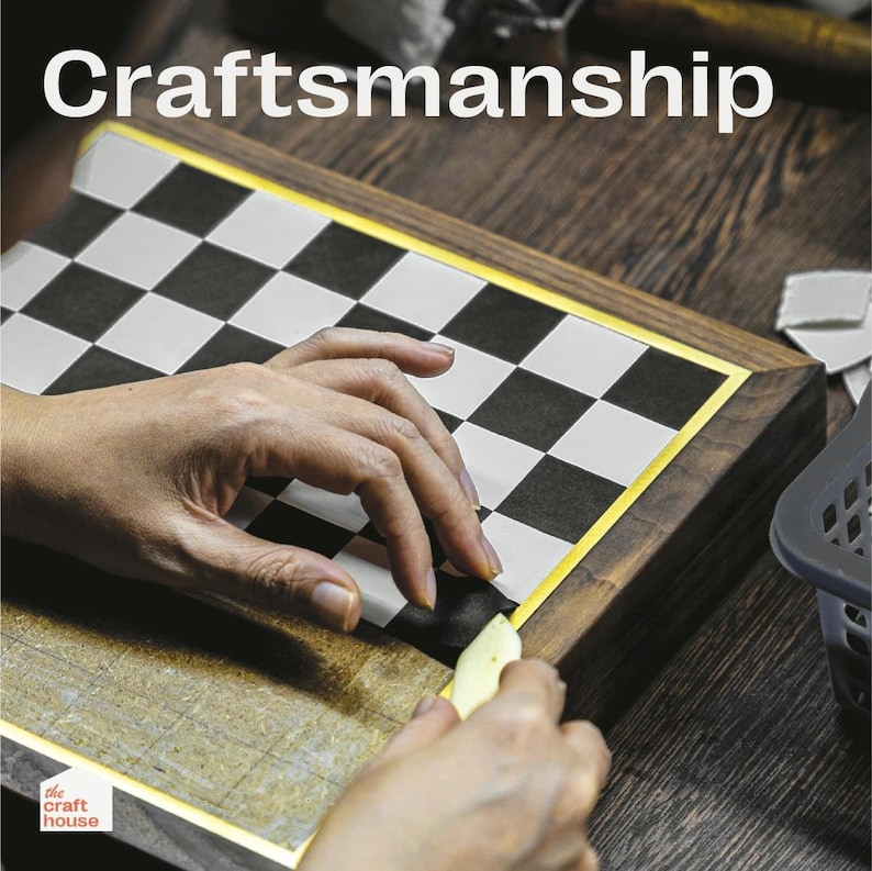Op de afbeelding: Een close-up van een schaakbord met zwart-witte vierkanten, omlijst door donker hout met een gouden rand. Het woord "Craftsmanship" staat bovenaan. Handen werken aan het bord.