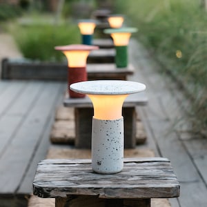 May include: A white outdoor lamp with a speckled finish and a round, flat top. The lamp is lit and casting a warm glow. The lamp is sitting on a wooden surface.