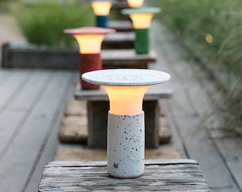 May include: A white outdoor lamp with a speckled finish and a round, flat top. The lamp is lit and casting a warm glow. The lamp is sitting on a wooden surface.