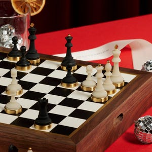 Op de afbeelding: Een schaakspel met zwarte en witte stukken op een schaakbord. De stukken zijn zwart en gebroken wit met goudkleurige bases. Het bord staat op een houten kist met een gouden rand. De achtergrond is rood.