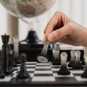 May include: A hand moves a clear glass chess piece on a black and white chessboard. The chessboard is surrounded by other chess pieces, including black and white pawns, rooks, and a king.