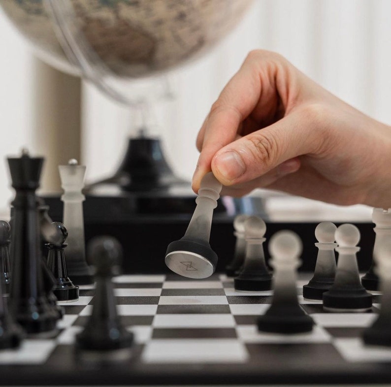 May include: A hand moves a clear glass chess piece on a black and white chessboard. The chessboard is surrounded by other chess pieces, including black and white pawns, rooks, and a king.