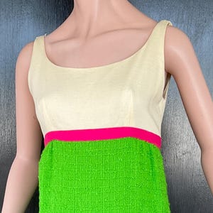 Pode incluir: Um vestido vintage sem mangas com um corpete creme e uma saia verde brilhante. Uma faixa rosa choque acentua a cintura e a bainha. O vestido é exibido em um manequim.