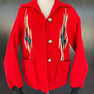 May include: A bright red long-sleeve jacket featuring a pointed collar, three silver-toned metal buttons, and two front pockets. The front panels display a vertical geometric pattern in shades of cream, black, and teal. The cuffs are black.