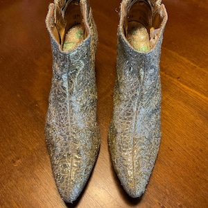 Könnte beinhalten: Ein Paar silber-goldene Stiefeletten mit spitzer Zehenpartie. Die Stiefel haben ein strukturiertes, gemustertes Design und einen goldenen Besatz am oberen Rand. Das Innere der Stiefel ist goldfarben mit grünen Akzenten.