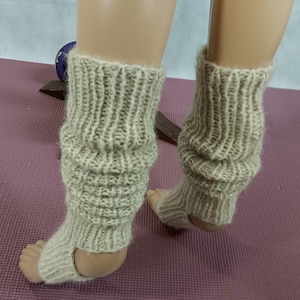 May include: A pair of beige knitted leg warmers with a ribbed design. The leg warmers have a foot opening and are displayed on a mannequin. The knitted texture is visible, and the color is a neutral beige.