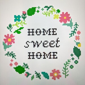 May include: A cross-stitch embroidery design featuring a wreath of green leaves and pink flowers with a yellow center. The wreath surrounds the text "Home Sweet Home".