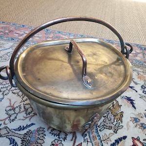 Puede incluir: Una olla de cobre antigua grande con tapa y asa de cobre. La olla tiene un cuerpo redondeado y una tapa ancha y plana. El asa está unida a la tapa y a la olla con dos lazos de cobre.