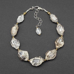May include: A necklace featuring large, faceted, clear quartz-like beads arranged in a circular pattern. The beads are separated by small, metallic gold and silver-toned spacers. It includes a silver-toned chain with a hook clasp and a single bead drop.