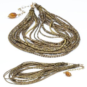 May include: A multi-strand necklace featuring numerous rows of metallic bronze-toned cylindrical and round beads. The design includes a gold-toned metal chain extender with a single amber-colored translucent bead at the end of the clasp mechanism.