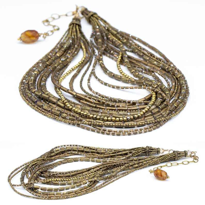 May include: A multi-strand necklace featuring numerous rows of metallic bronze-toned cylindrical and round beads. The design includes a gold-toned metal chain extender with a single amber-colored translucent bead at the end of the clasp mechanism.