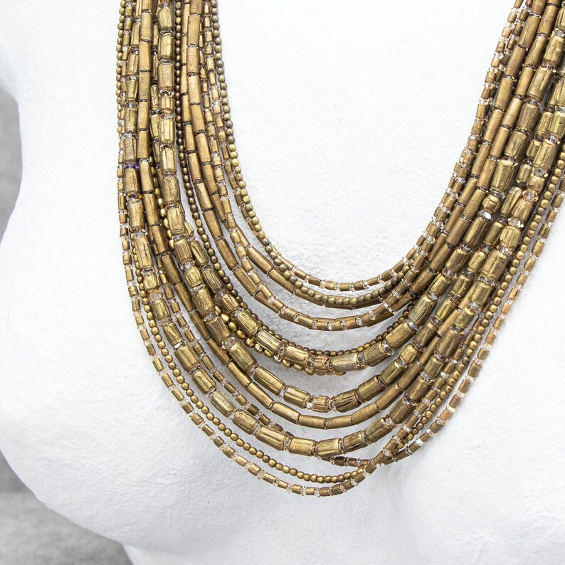 May include: A multi-strand necklace featuring numerous layers of antique gold-toned cylindrical beads and small round metallic beads. The strands are arranged in a cascading, graduated design, creating a textured and layered appearance.