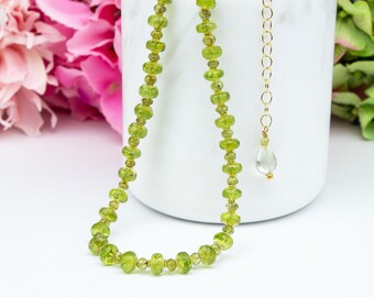 May include: A necklace featuring faceted lime green peridot beads strung on a thin gold-toned wire. The necklace includes a gold-toned metal chain extender with a small, clear, teardrop-shaped pendant attached to the end of the adjustable clasp.