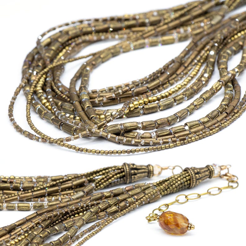 May include: A multi-strand necklace featuring numerous antique brass-toned cylindrical beads interspersed with small clear faceted crystals and tiny metallic spheres. The necklace includes a gold-toned chain extender with a single amber-colored bead.