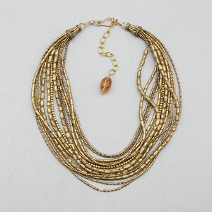 May include: A multi-strand necklace featuring numerous rows of metallic gold-toned cylindrical and round beads. The necklace includes a gold-toned metal hook clasp, an adjustable chain, and a single amber-colored faceted bead at the end.