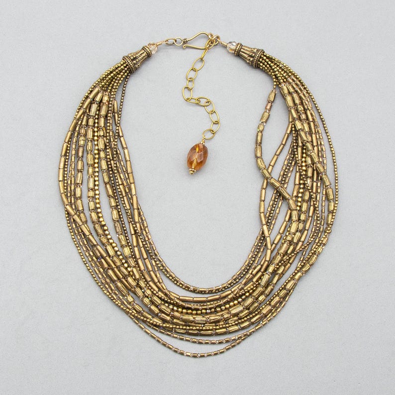 May include: A multi-strand necklace featuring numerous rows of metallic gold-toned cylindrical and round beads. The necklace includes a gold-toned metal hook clasp, an adjustable chain, and a single amber-colored faceted bead at the end.