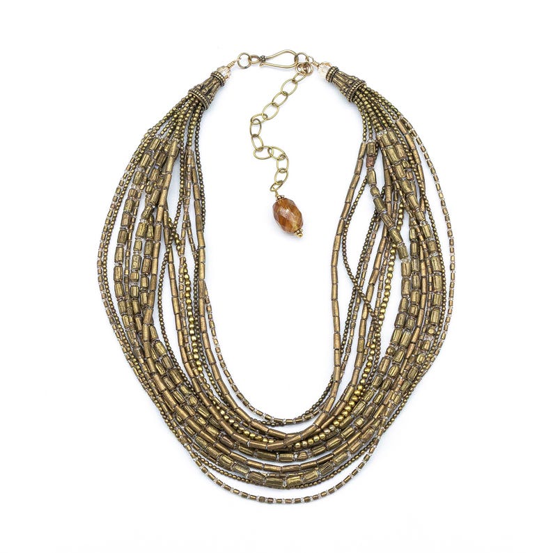 May include: A multi-strand necklace featuring numerous rows of antique brass-toned cylindrical beads and small metallic spheres. The piece includes a metal hook clasp, an adjustable chain extension, and a single faceted amber-colored bead pendant.