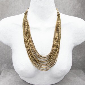 May include: A multi-strand necklace featuring numerous rows of metallic gold-toned cylindrical and spherical beads. The strands are gathered at the top with ornate gold-toned caps, creating a cascading, layered effect that drapes down the chest.
