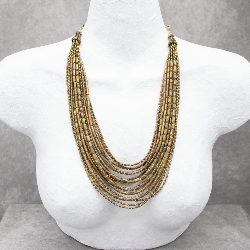 May include: A multi-strand necklace featuring numerous rows of metallic gold-toned cylindrical and spherical beads. The strands are gathered at the top with ornate gold-toned caps, creating a cascading, layered effect that drapes down the chest.