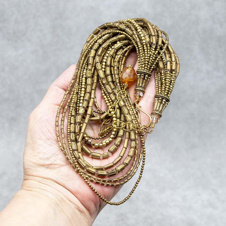 May include: A multi-strand necklace featuring numerous rows of metallic bronze-toned cylindrical and round beads. The design includes a single amber-colored faceted stone accent, a gold-toned metal clasp, and an adjustable chain link extender.