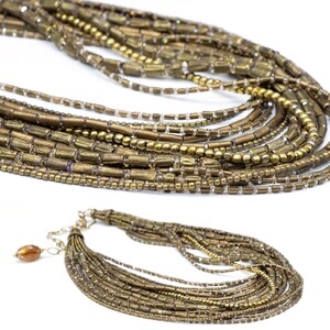 May include: A multi-strand necklace featuring numerous rows of metallic bronze-toned tube beads and small round beads interspersed with clear, reflective glass seed beads. The necklace includes a metal chain extender and a single amber-colored bead.