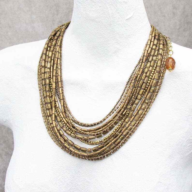 May include: A multi-strand necklace featuring numerous rows of metallic bronze-toned cylindrical and round beads. The layered design includes a gold-toned chain extension at the side, finished with a single translucent amber-colored faceted bead.