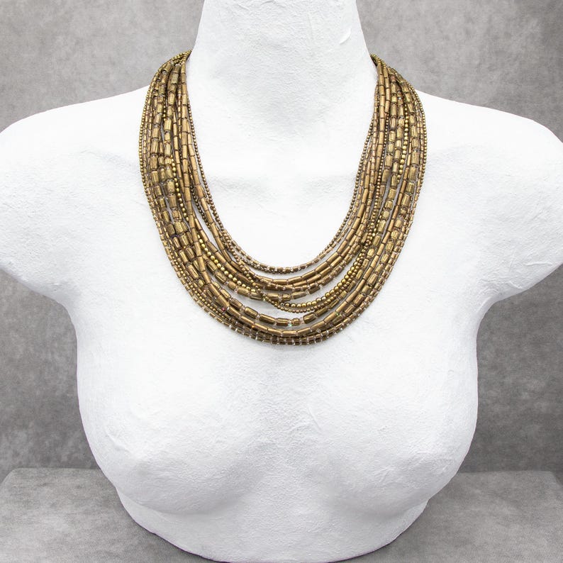 May include: A multi-strand necklace featuring several layers of metallic bronze-toned cylindrical beads and small round beads. The necklace is arranged in a cascading, graduated design that rests around the neck, showcasing a textured finish.